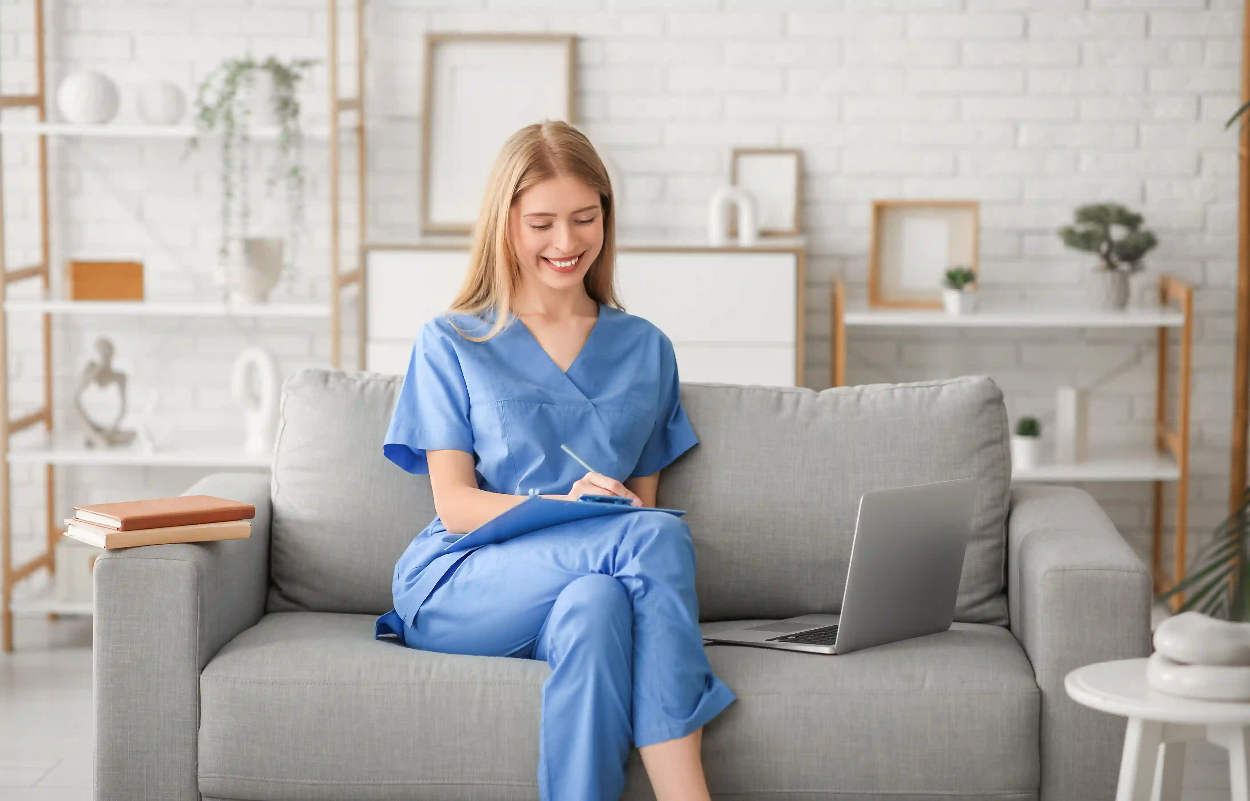 Travel nurse in blue scrubs planning her move in a furnished apartment with a laptop and clipboard.