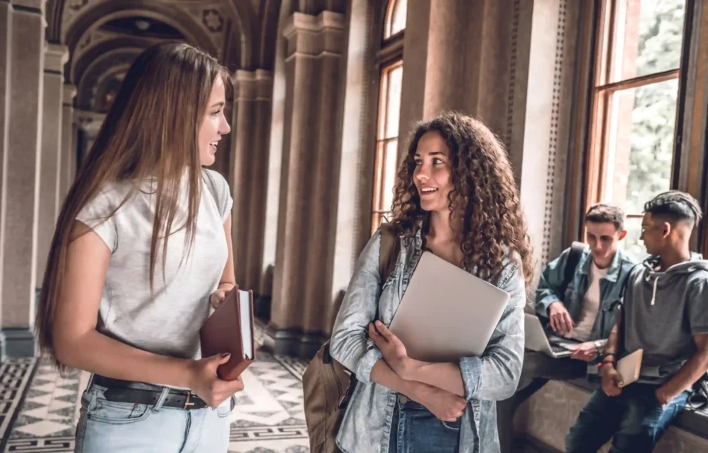 Moving with teenagers as students talk in a school hallway while holding books and a laptop.