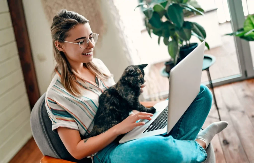 Woman working on a laptop with a cat on her lap while planning pet transport at home.