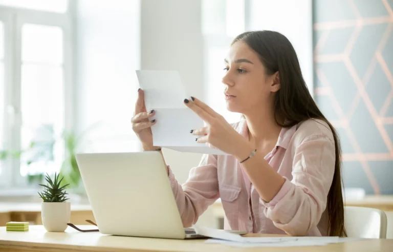 Young woman reviewing an employee relocation letter beside a laptop in a bright office.